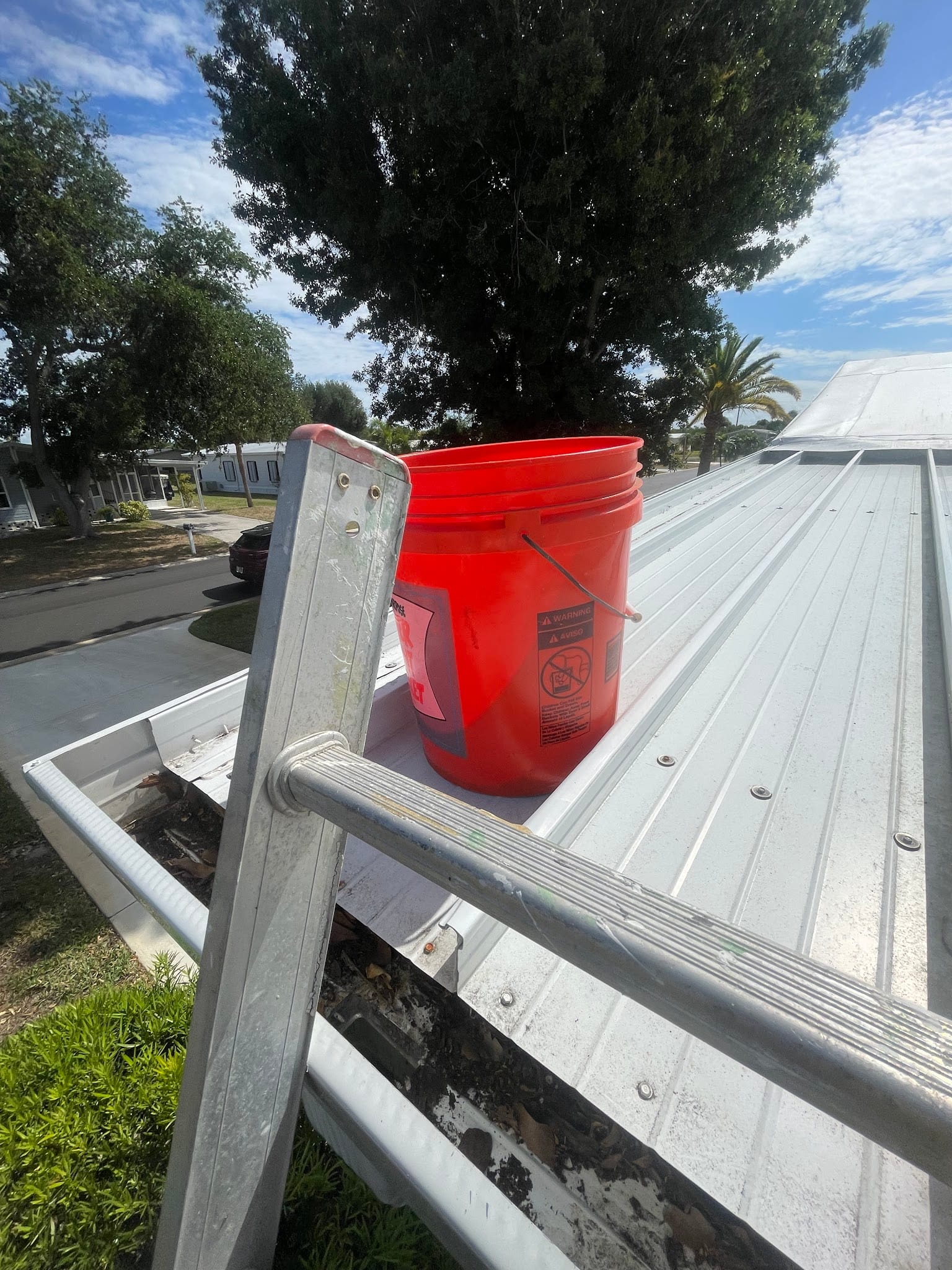 Red 5-gallon bucket on metal roof next to ladder during gutter cleaning